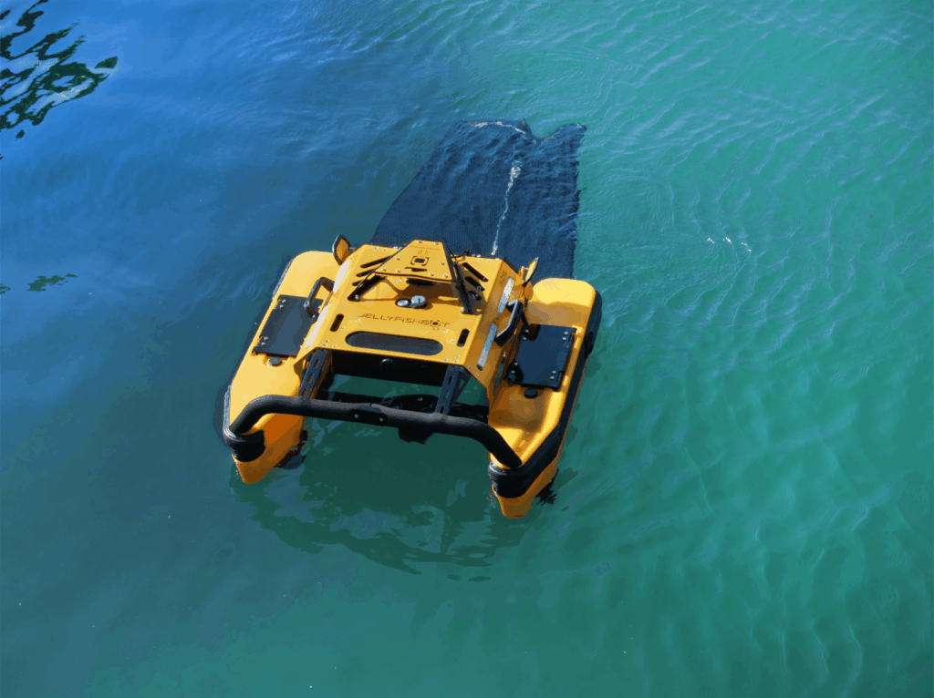 Photo of a yellow Jellyfishbot in blue water; it's pulling a net to collect waste from the water.