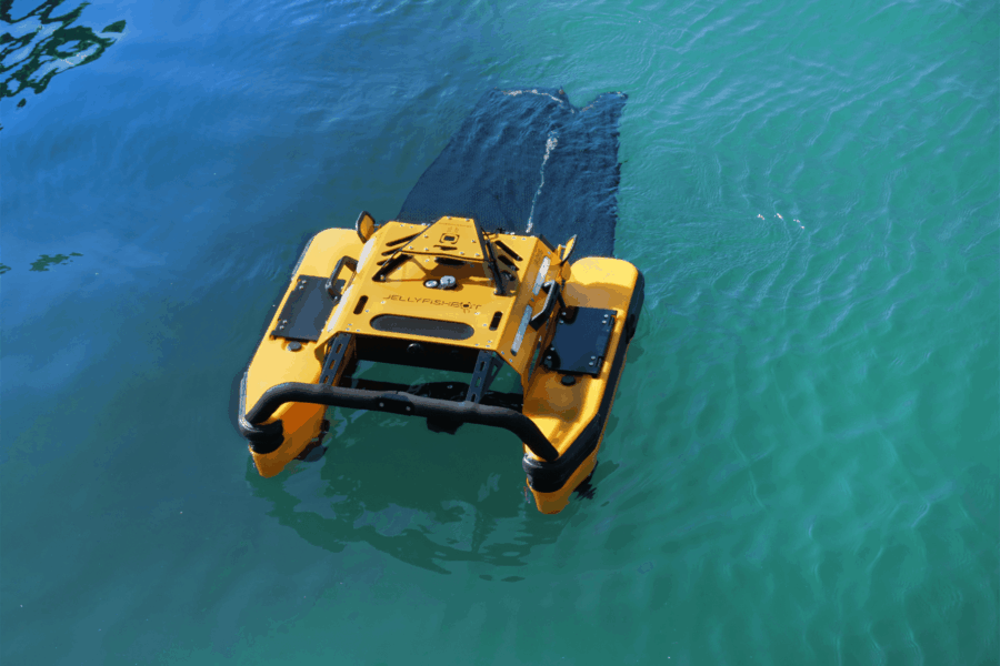Photo of a yellow Jellyfishbot in blue water; it's pulling a net to collect waste from the water.